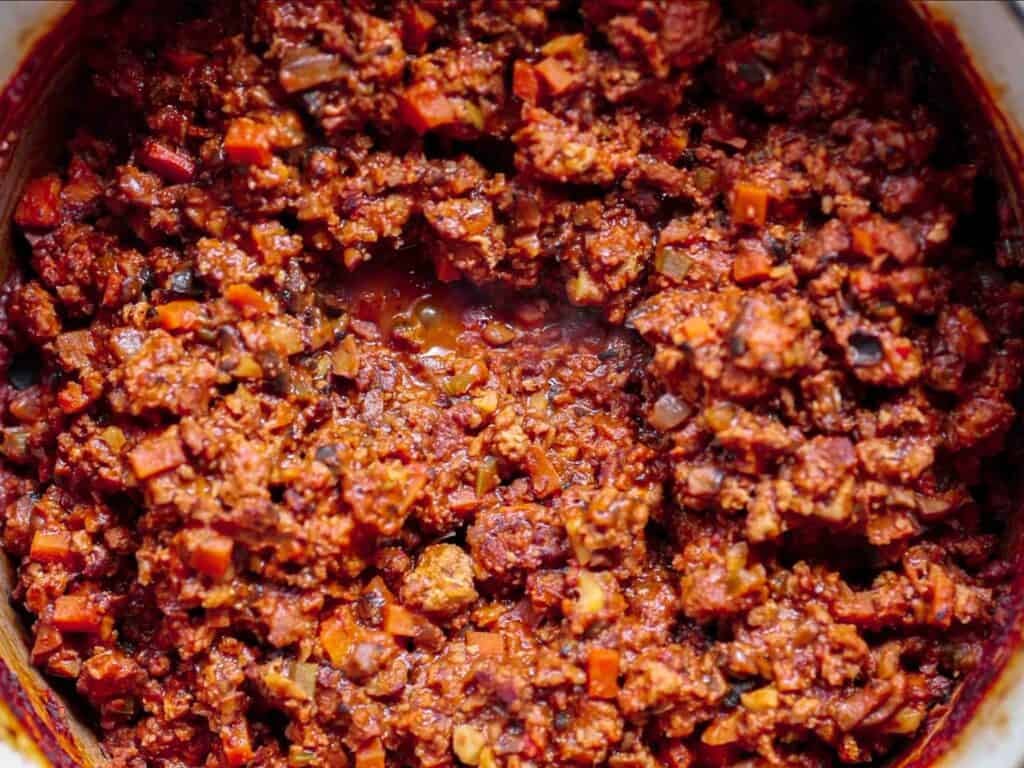 Close-up of a pot filled with chunky, cooked vegan bolognese sauce featuring plant-based ground meat, diced carrots, onions, and tomato sauce. The mixture has a rich red-brown color and a hearty texture.